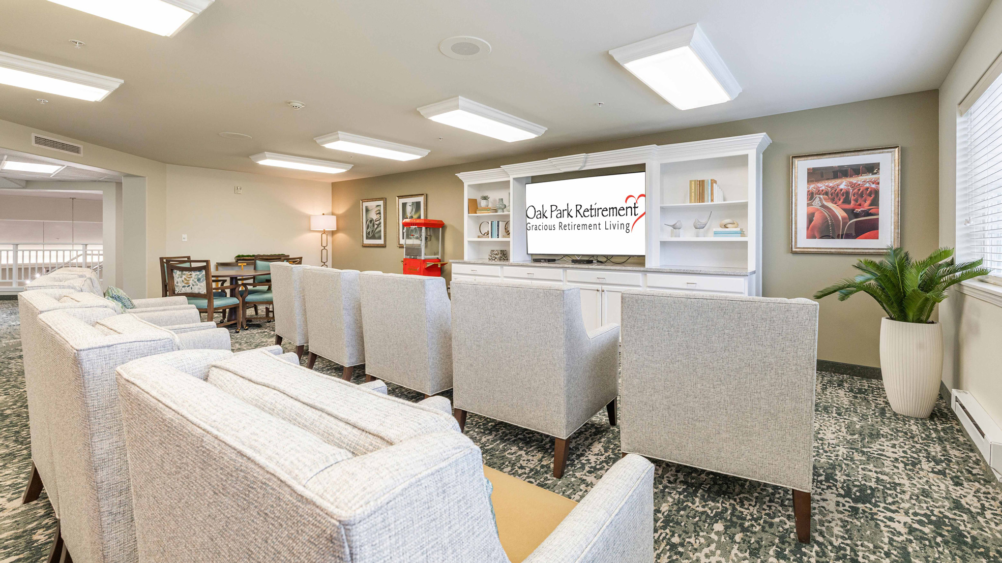 A bright, modern lounge area with cushioned chairs and sofas facing a TV displaying "Oak Park Retirement." Shelves with decor and books, a table with chairs, artwork, and large windows complete the inviting space.
