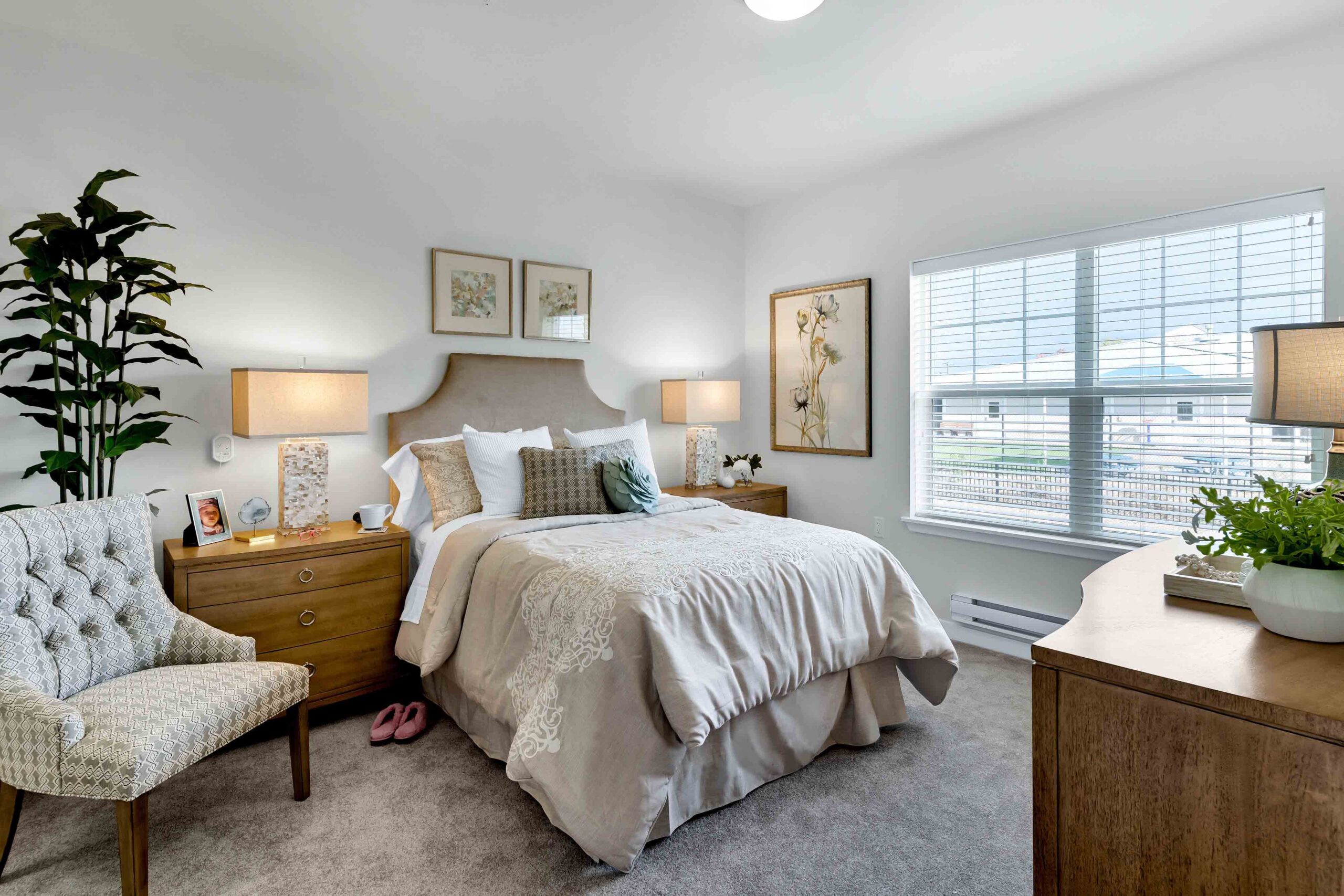 A cozy bedroom features a bed with beige bedding and decorative pillows, flanked by wooden nightstands with lamps. A small cushioned chair, dresser, large potted plants, and framed artwork adorn the space. A large window fills the room with natural light.