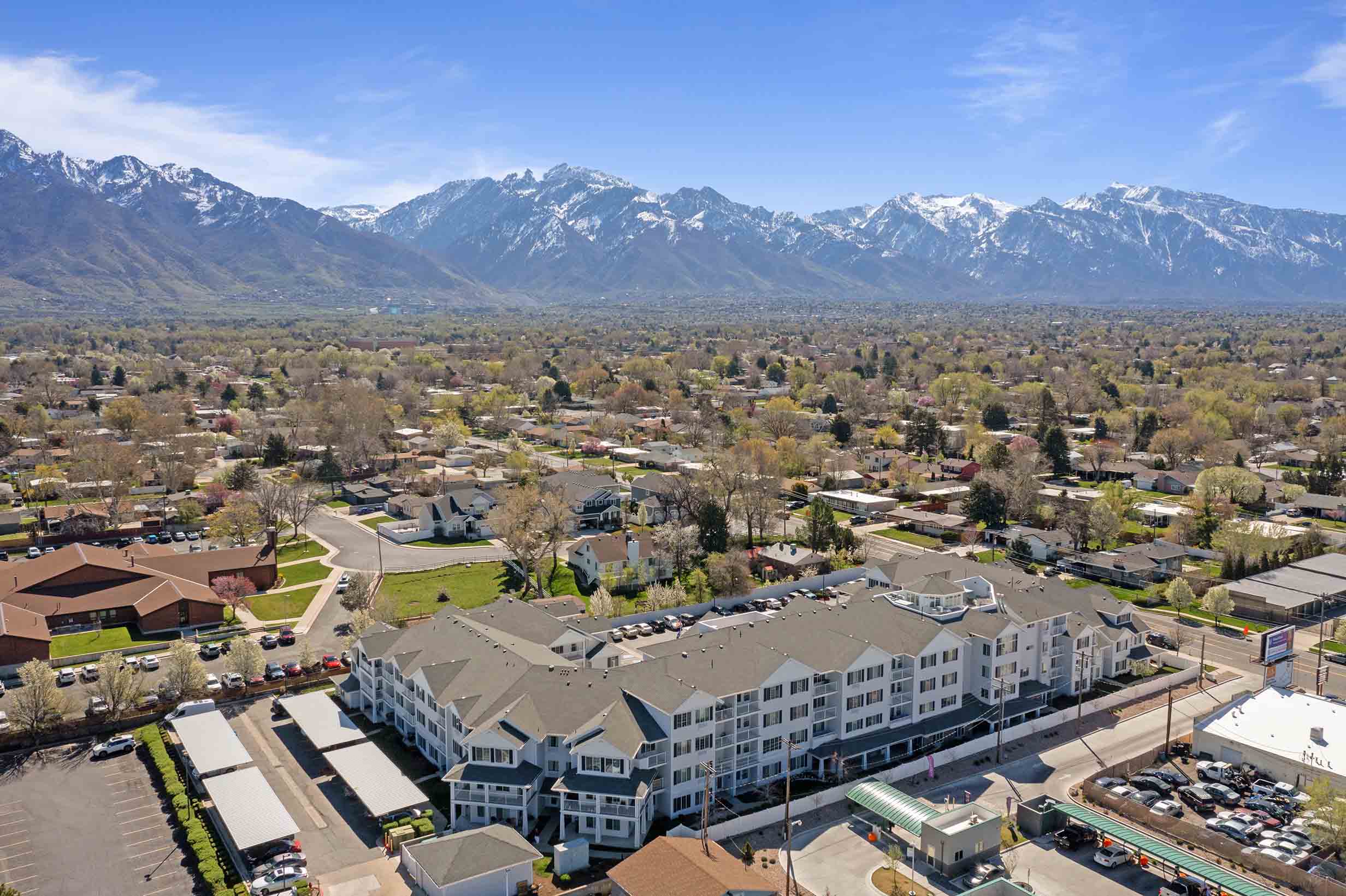 Aerial view of a suburban neighborhood with residential buildings surrounded by greenery. In the background, there are majestic snow-capped mountains under a clear blue sky. The foreground features a large building complex and streets lined with parked cars.