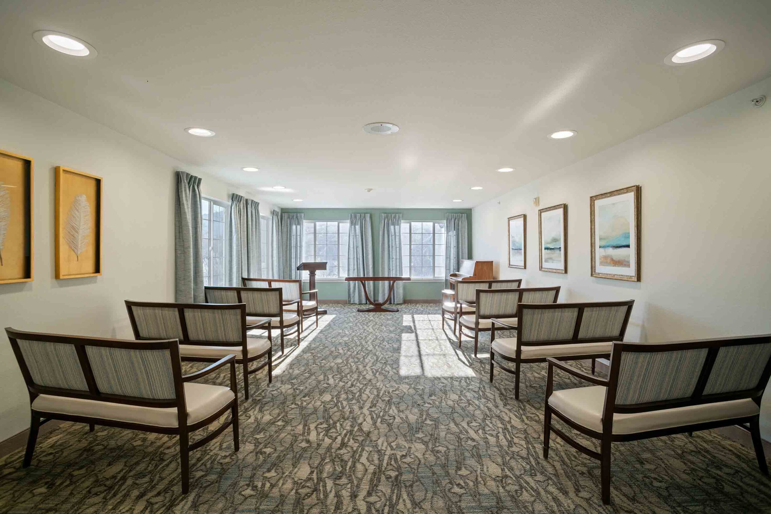 A well-lit room with several wooden chairs arranged in rows facing a podium and window. The floor is carpeted, and the walls are adorned with framed artwork. The large windows at the back let in natural light, and there are gray curtains on either side.