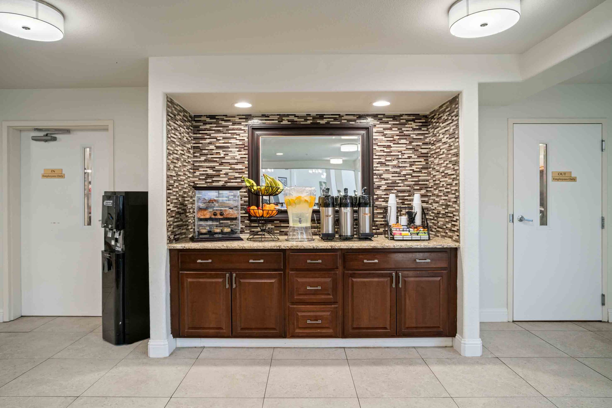 A well-lit refreshment area featuring a dark wood cabinet stocked with coffee dispensers, tea bags, and fresh fruit. Above the counter is a large mirror framed by mosaic tiles. To the left is a water dispenser, and on the right are doors labeled 