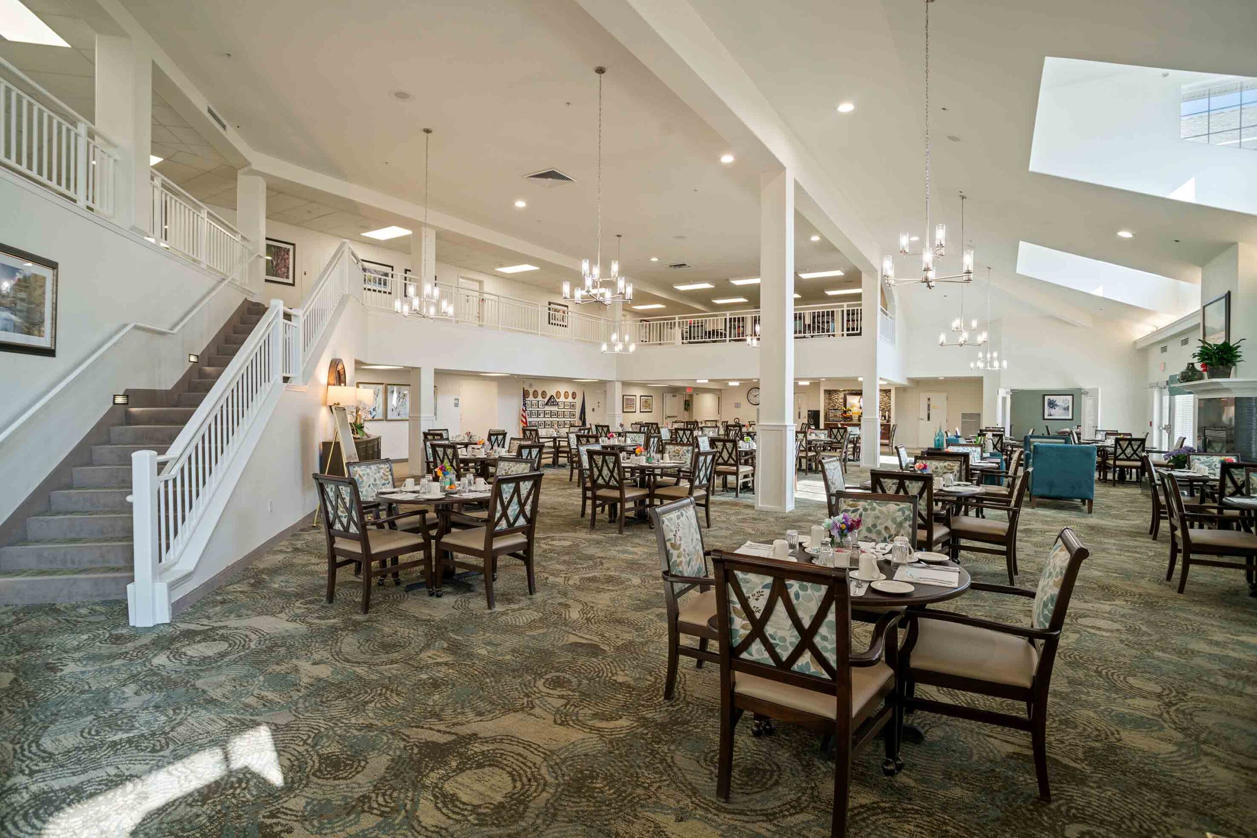 A spacious, well-lit dining area with tables and chairs arranged neatly. There is a staircase to the left leading to an upper level with a railing. Chandeliers hang from the high ceiling, and the room has large windows allowing natural light to flood in.