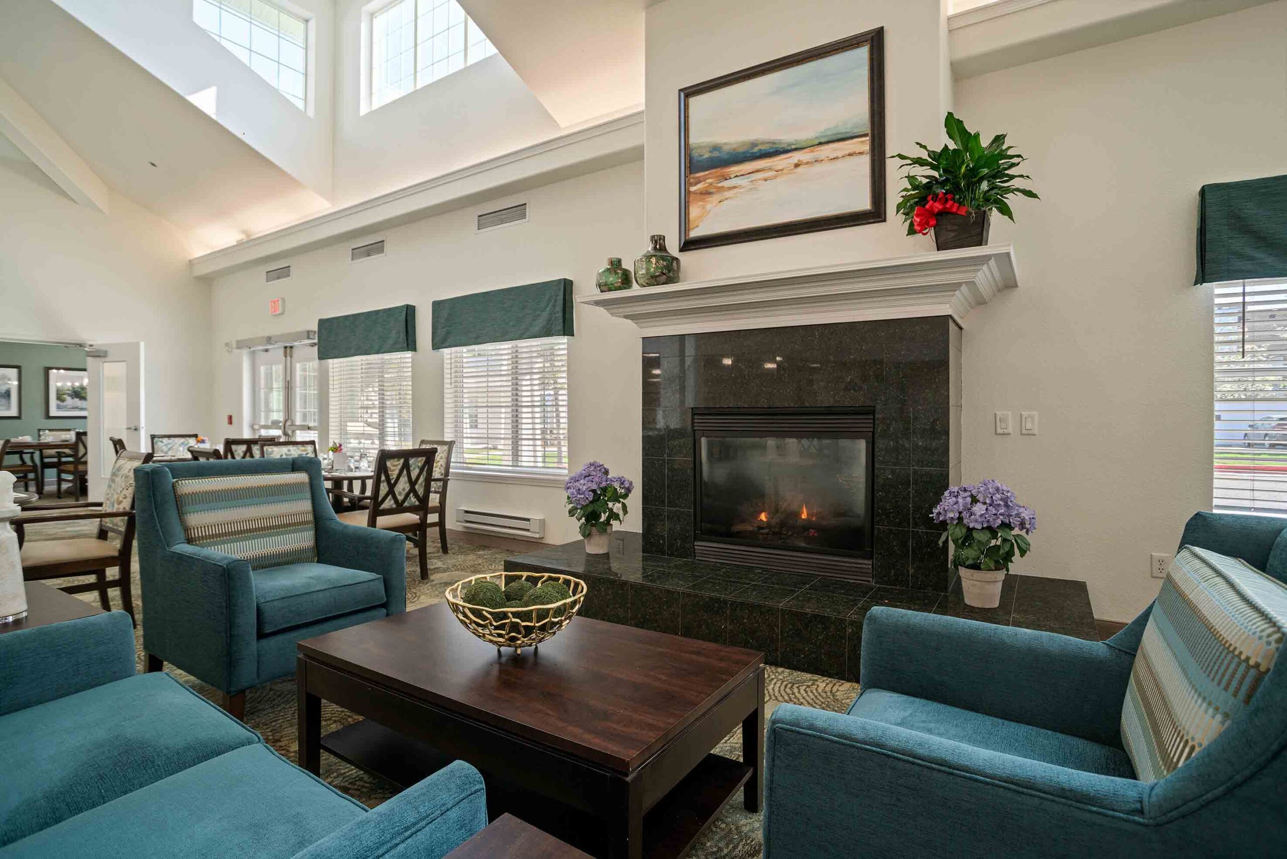 A cozy living room featuring a blazing fireplace framed by dark stone. Two blue armchairs, a wooden coffee table with a decorative bowl, and a green plant adorn the space. Large windows and high ceilings allow ample natural light. Dining tables are visible in the background.