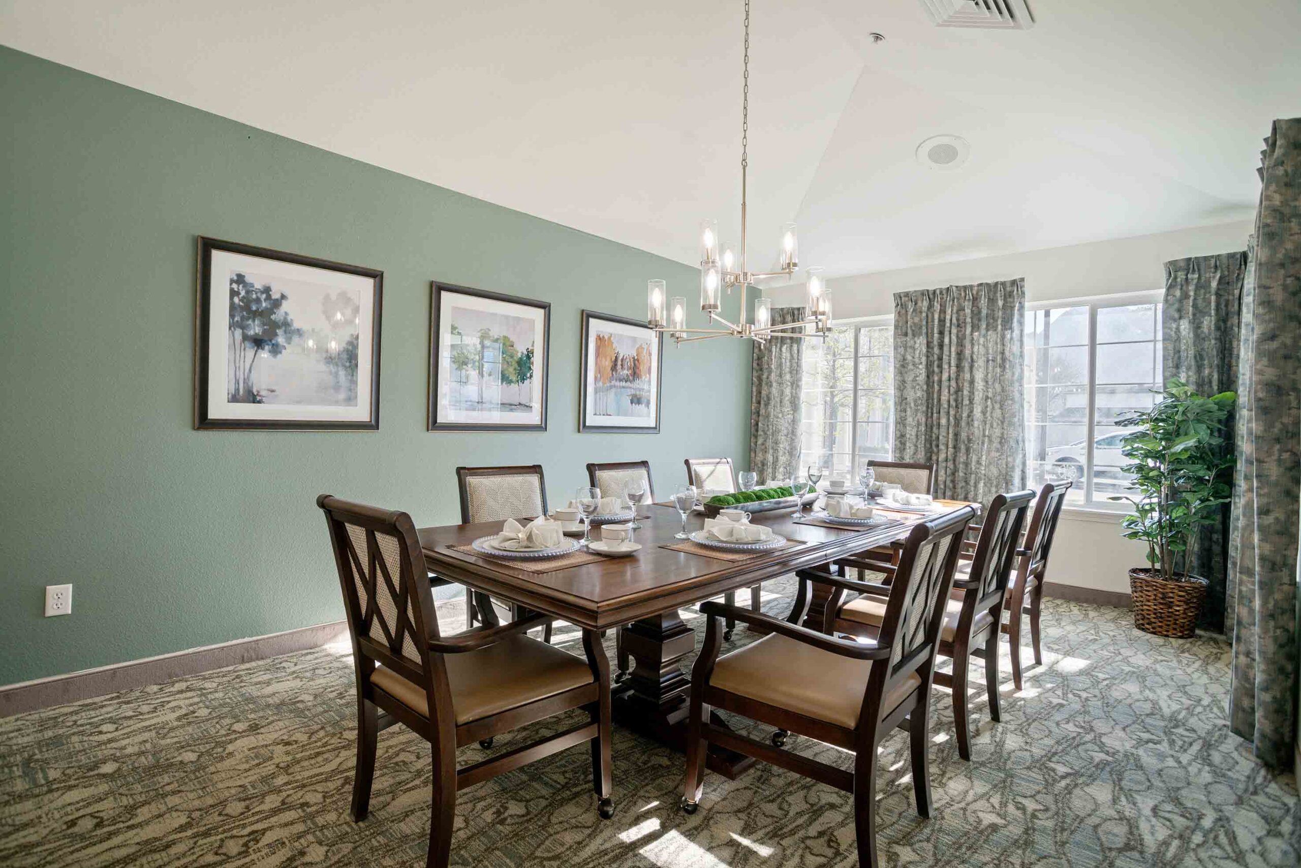 A dining room with a long wooden table set for eight, featuring cushioned chairs. A modern chandelier hangs above. The room has light green walls with framed artwork and large windows with patterned curtains letting in natural light. A potted plant is in the corner.