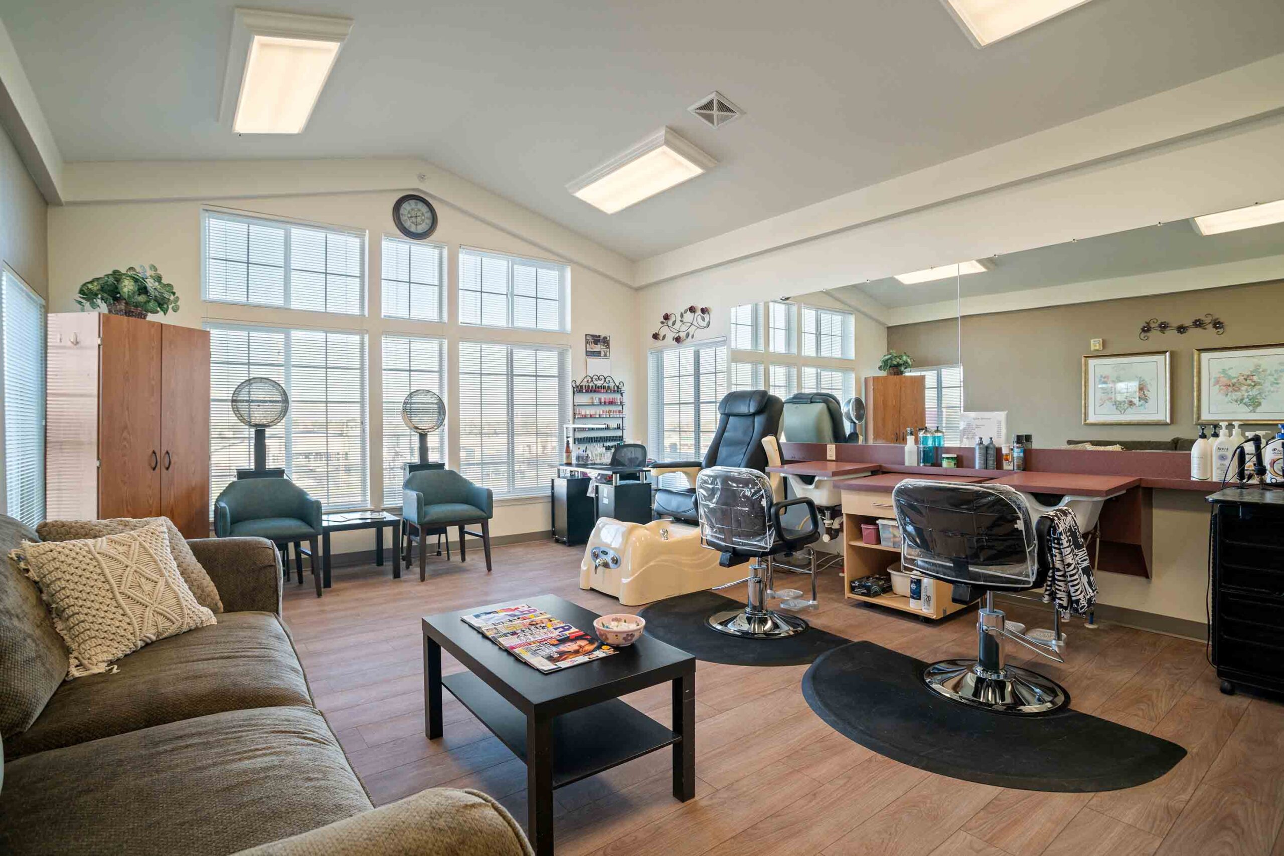 A well-lit salon room featuring two styling chairs in front of a large mirror and a counter with hair products. A dryer station with two hooded hair dryers is against the window. The space includes a sofa, chairs, magazines on a coffee table, and hardwood floors.