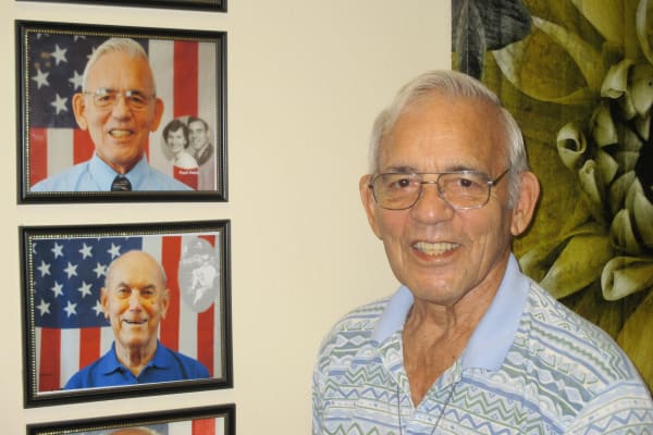 An elderly man with glasses and wearing a patterned shirt smiles at the camera. Behind him, two framed photos show men in front of American flags. One photo includes a grayscale image of a couple in the background.