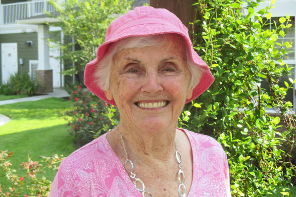 An elderly woman with short white hair smiles warmly. She is wearing a pink hat and a pink patterned top, accessorized with a silver necklace. She is standing outdoors in a lush green setting with plants and a building in the background.