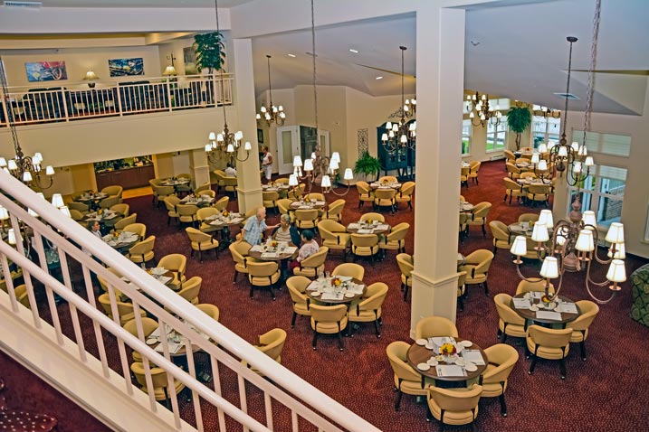 A spacious, well-lit restaurant with high ceilings and multiple chandeliers. The room is filled with round tables and beige chairs, with several patrons dining. A mezzanine area is visible on the left, overlooking the main dining area. Red carpeting covers the floor.