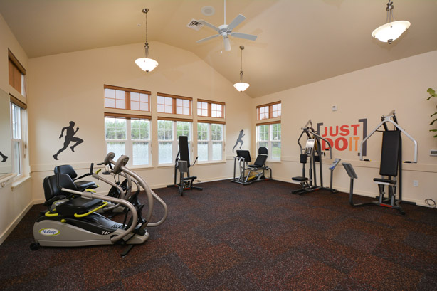 A spacious gym with large windows on three walls. It features various exercise equipment, including treadmills, exercise bikes, and weight machines. The walls are decorated with silhouette decals of athletes and the slogan 