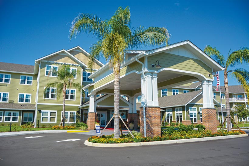 A modern, three-story building with a green and beige exterior features a large covered entrance and brick columns. Palm trees and landscaped gardens surround the entrance. The sky is clear and blue, creating a welcoming and vibrant atmosphere.