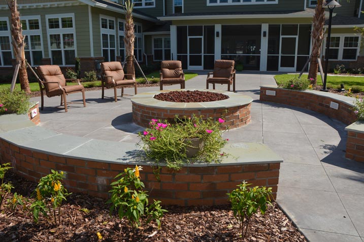 A circular outdoor seating area features a central fire pit surrounded by cushioned chairs. Raised brick planters with blooming plants encircle the fire pit. The backdrop is a building with large windows and greenery. The patio is sunlit.