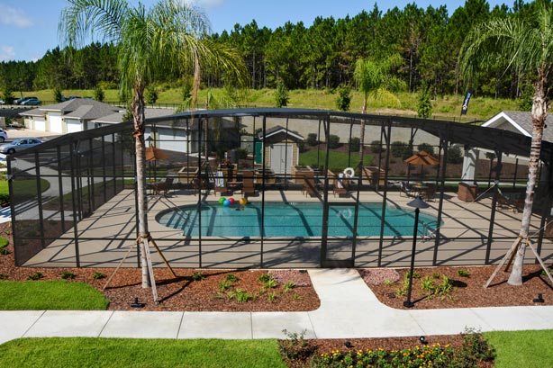 A backyard pool is enclosed in a large black screen, surrounded by a concrete deck and landscaped garden. Taller trees and shrubs line the outside, while colorful pool toys float in the water. In the background, a house and wooded area can be seen.