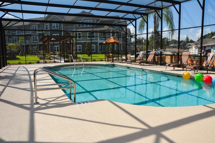 Outdoor swimming pool enclosed in a screened structure, surrounded by lounge chairs, umbrellas, and three colorful beach balls floating on the water. In the background, there are apartment buildings and landscaped greenery. The sky is clear and sunny.