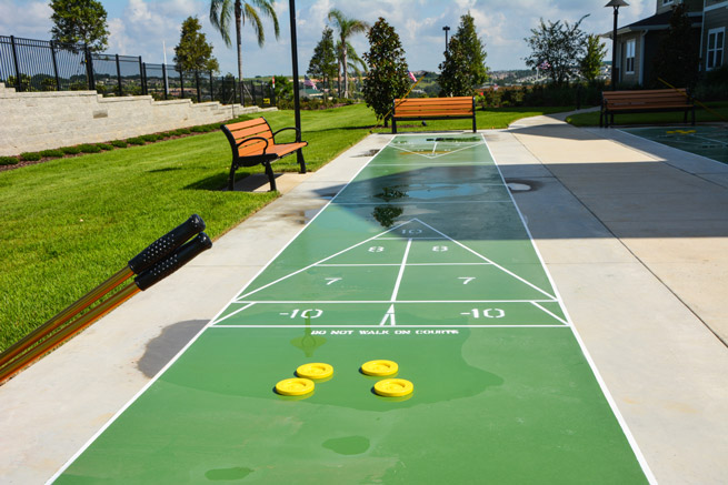 A sunny outdoor shuffleboard court with four yellow pucks placed randomly on the green playing surface. The court is surrounded by benches, grass, and trees, with a clear blue sky in the background.