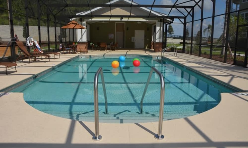 A screened-in, rectangular swimming pool with three colorful inflatable balls floating on the water. Poolside lounge chairs, an umbrella, and a lifebuoy are visible on the left. The background shows a small building with large glass doors and a clear sky above.