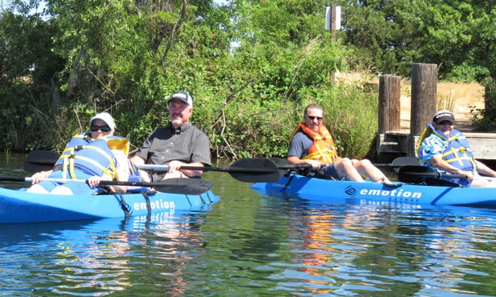 Three men are sitting in blue tandem kayaks on a calm body of water. They are wearing life jackets and sunglasses. One is wearing a hat, another has a cap, and the third has short hair. There are green trees and foliage in the background, along with a wooden dock.