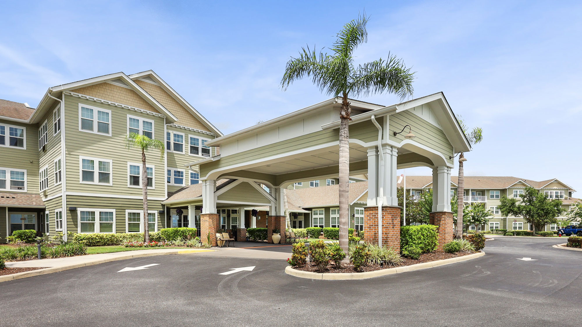 A modern, multi-story senior living facility with a covered driveway entrance, surrounded by palm trees and landscaping under a clear blue sky.
