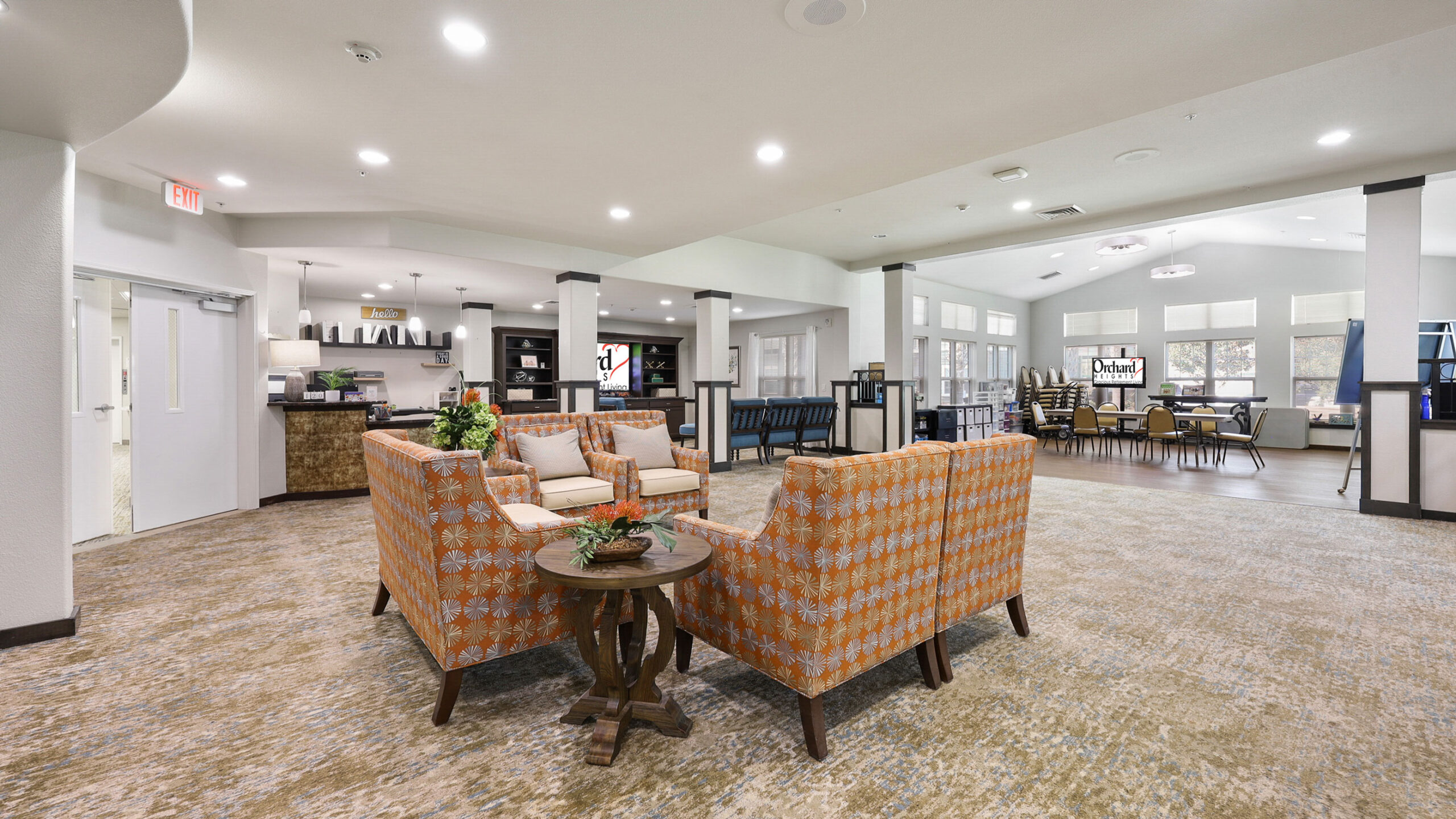 A spacious, well-lit lounge area with patterned armchairs around a small table, neutral carpeting, and a dining area with stacked chairs in the background. Large windows let in natural light.