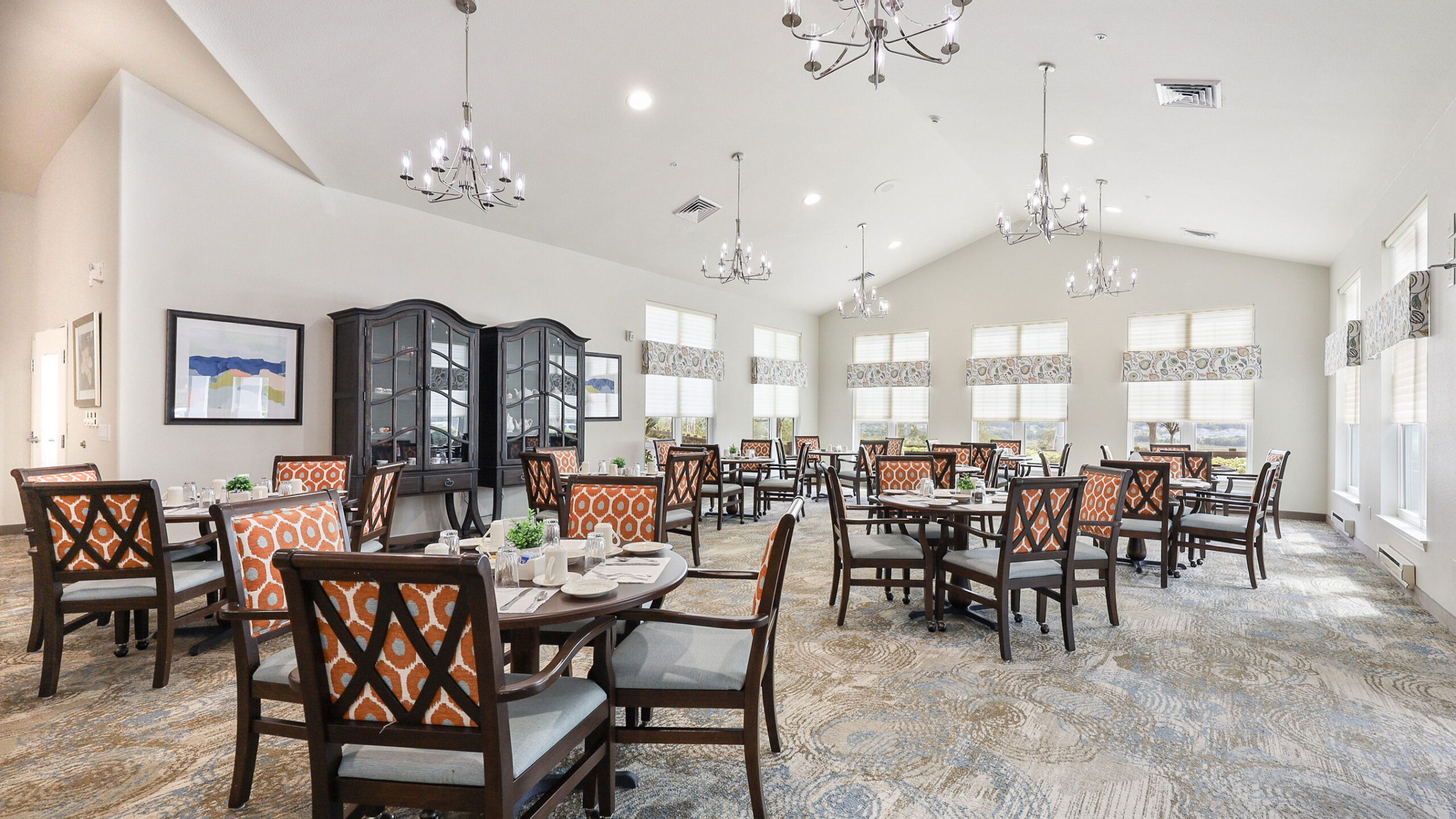 A bright, spacious dining room with multiple tables set for meals, orange-patterned chairs, large windows, white walls, modern chandeliers, and framed art on the walls.