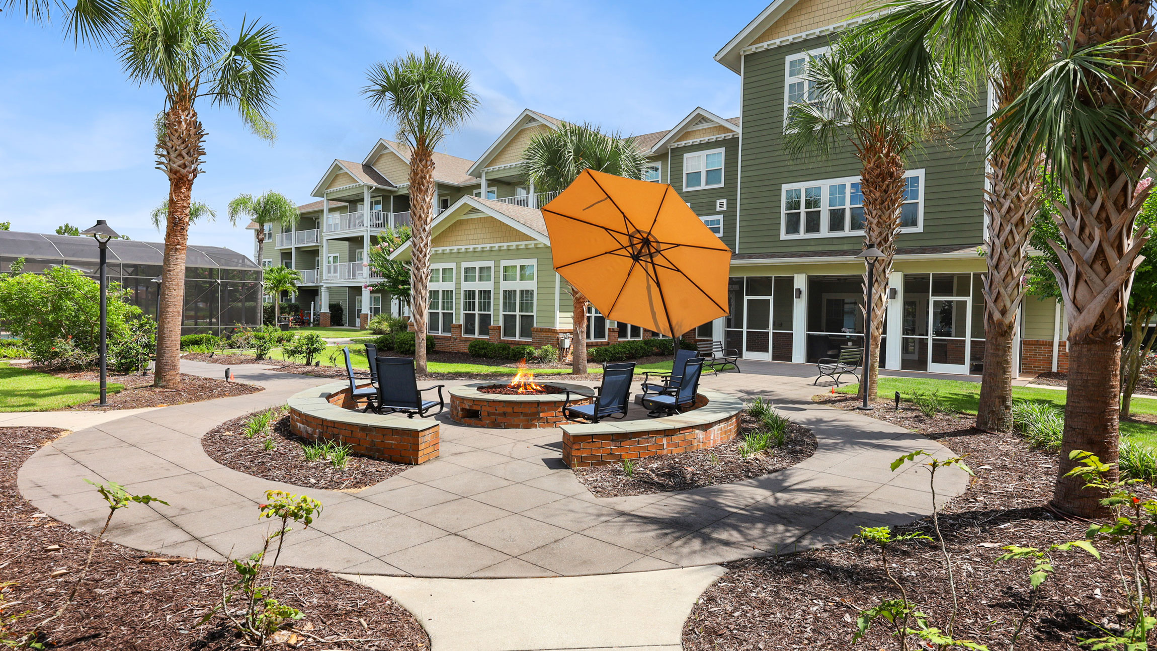 A landscaped courtyard with palm trees, a circular fire pit surrounded by chairs, and a large orange umbrella, in front of a modern apartment building under a clear blue sky.