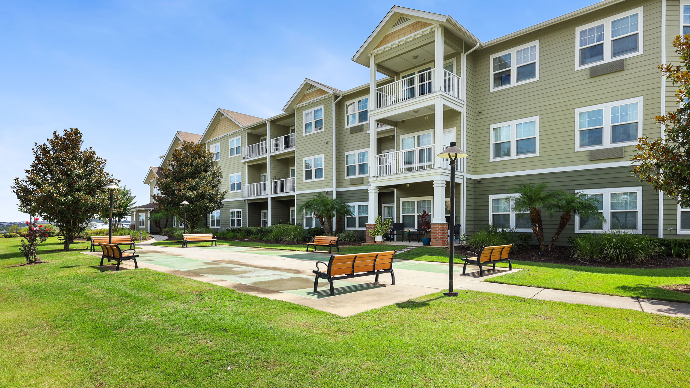 A modern three-story apartment building with balconies, surrounded by green grass, trees, and benches along a wide walkway on a sunny day.