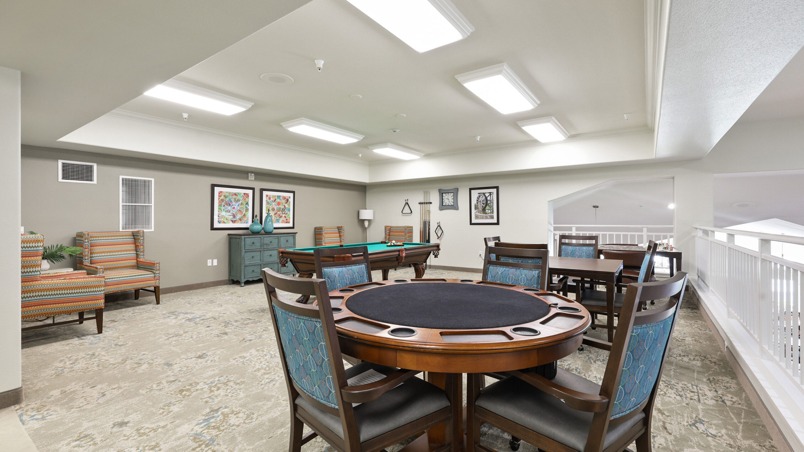 A bright, modern game room with poker tables, upholstered chairs, framed art on the walls, and patterned carpet. Overhead fluorescent lights illuminate the spacious area.