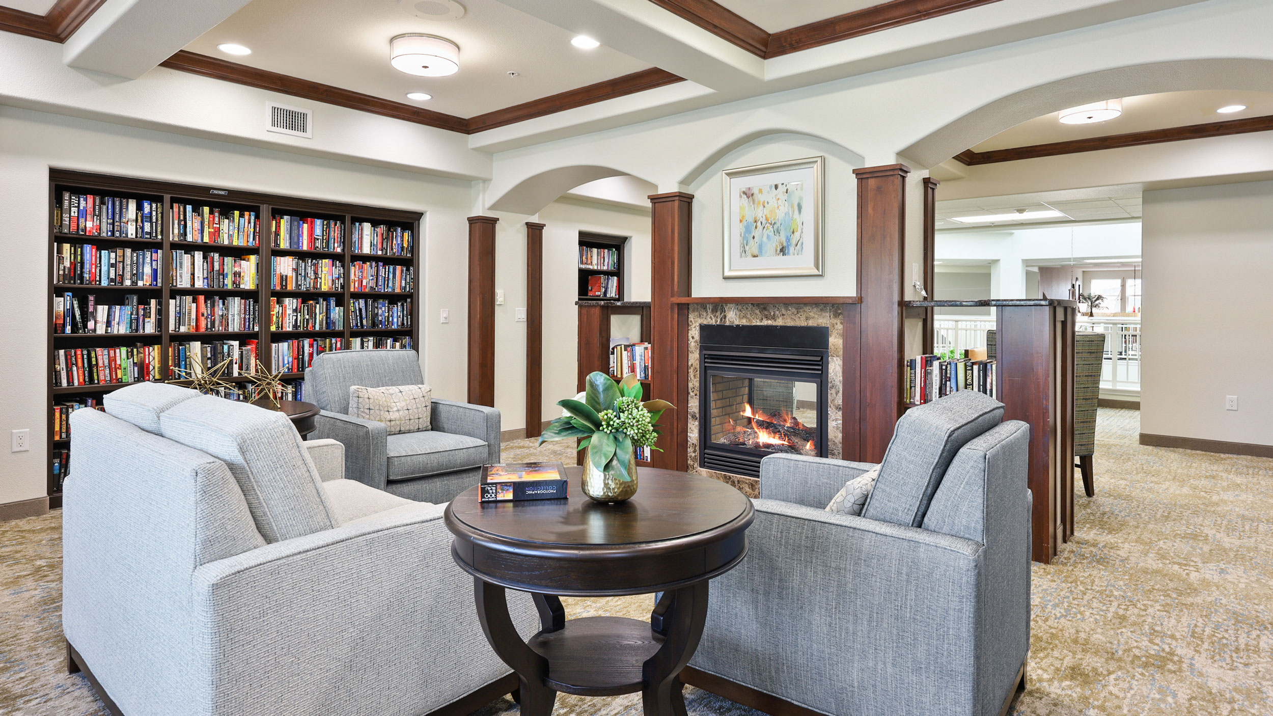 A cozy library with four gray armchairs around a small round table, a fireplace with a painting above it, built-in bookshelves filled with books, and soft lighting from ceiling fixtures.