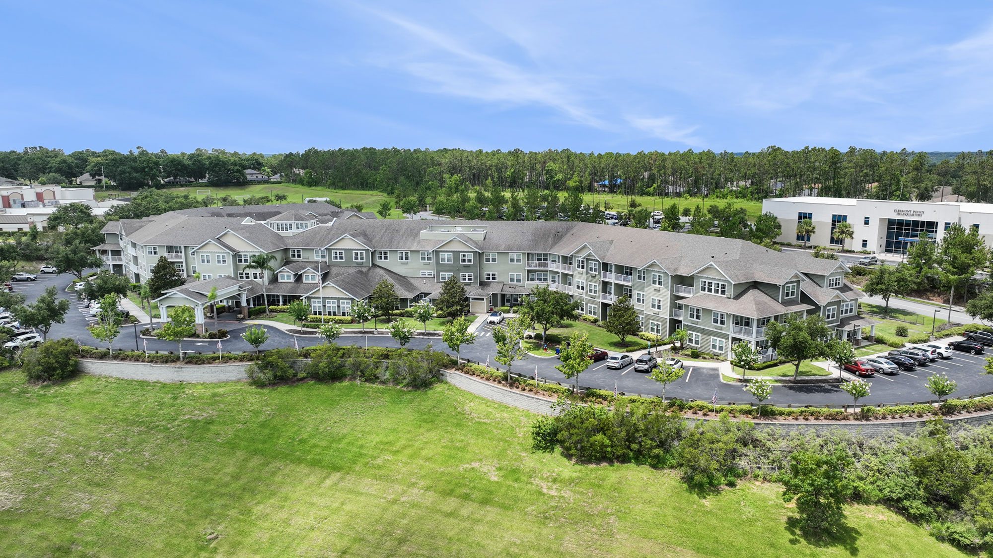 Aerial view of a large, multi-story building complex surrounded by trees, a parking lot with cars, and well-maintained green lawns under a blue sky.
