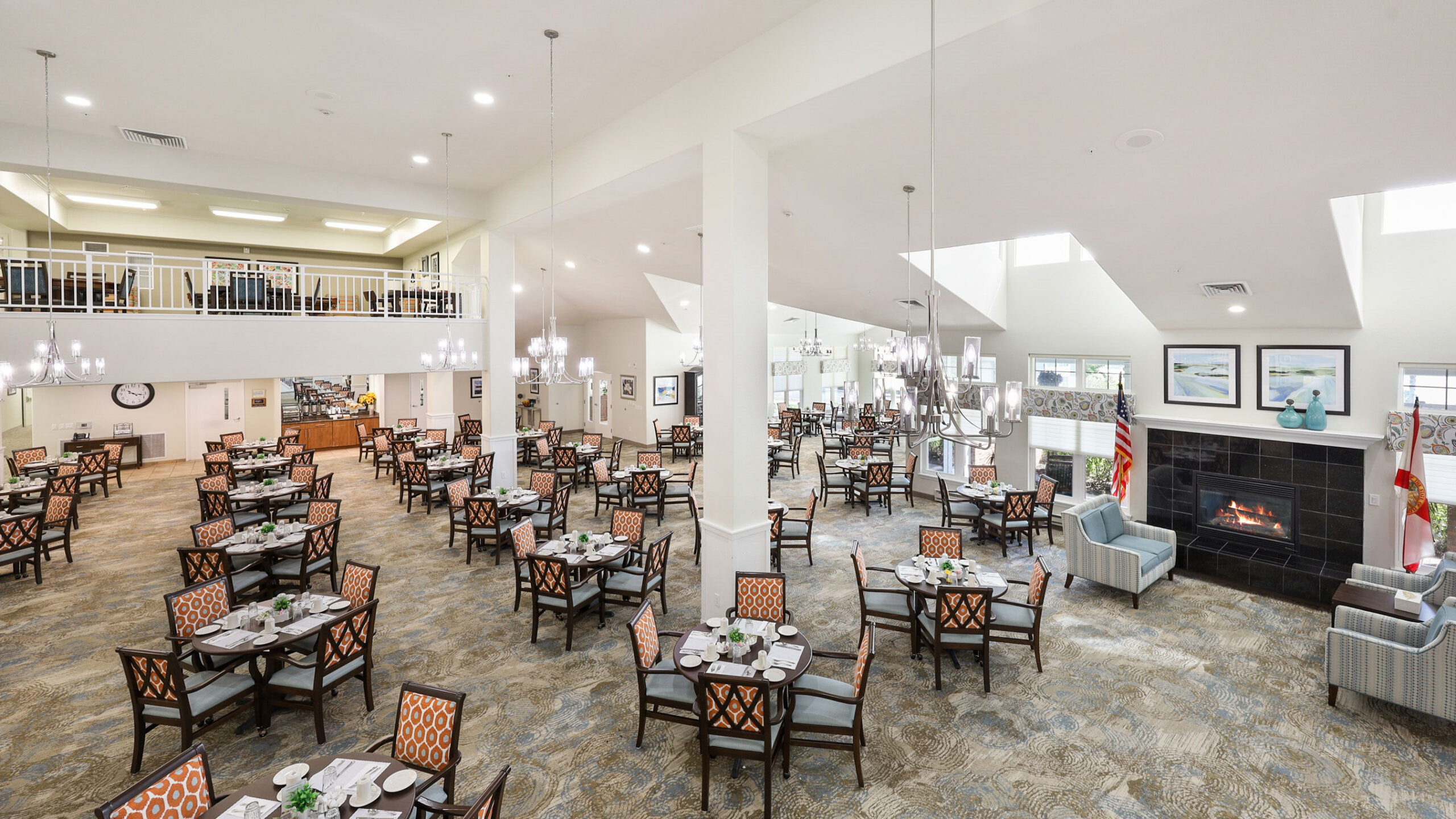 Spacious, well-lit dining hall with many set tables and patterned chairs, chandeliers, large windows, a fireplace, wall art, and American flags. A balcony overlooks the main dining area.