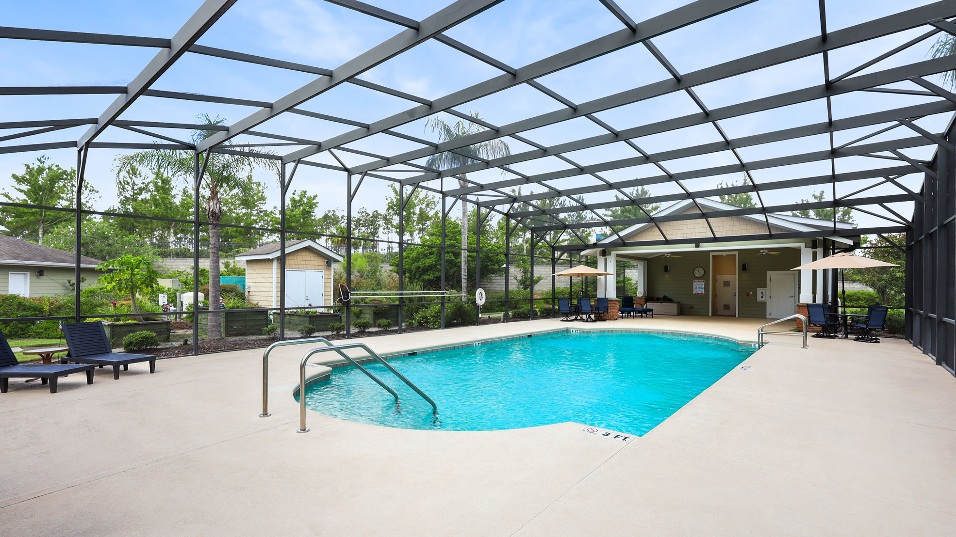 Enclosed outdoor swimming pool with clear blue water, surrounded by lounge chairs and umbrellas. The area is covered by a black metal screen structure with a small building and greenery in the background.