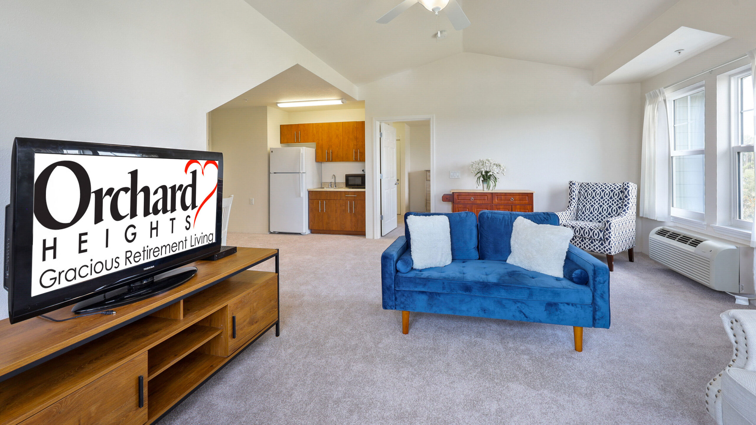 A bright living room with a blue couch, TV displaying "Orchard Heights Gracious Retirement Living," wooden furniture, kitchenette, and large windows letting in natural light.