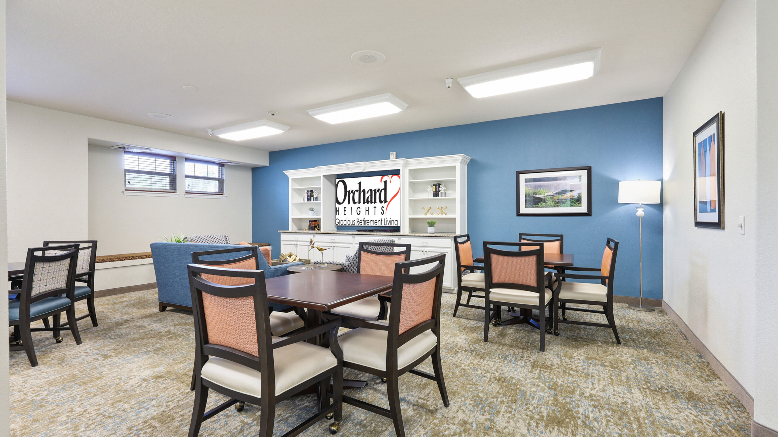 A brightly lit common room with several tables and chairs, a blue accent wall, artwork, and a white entertainment center displaying a sign that reads "Orchard Heights.