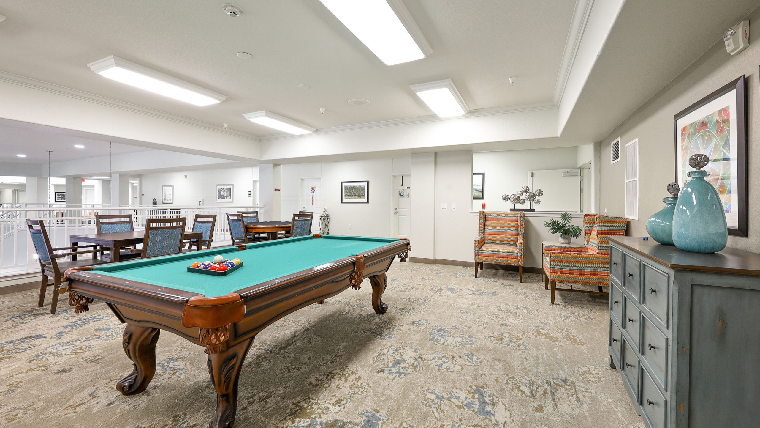A well-lit recreation room with a pool table, cue sticks, two striped armchairs, a blue sideboard with decorative vases, and a wooden dining table with chairs in the background.