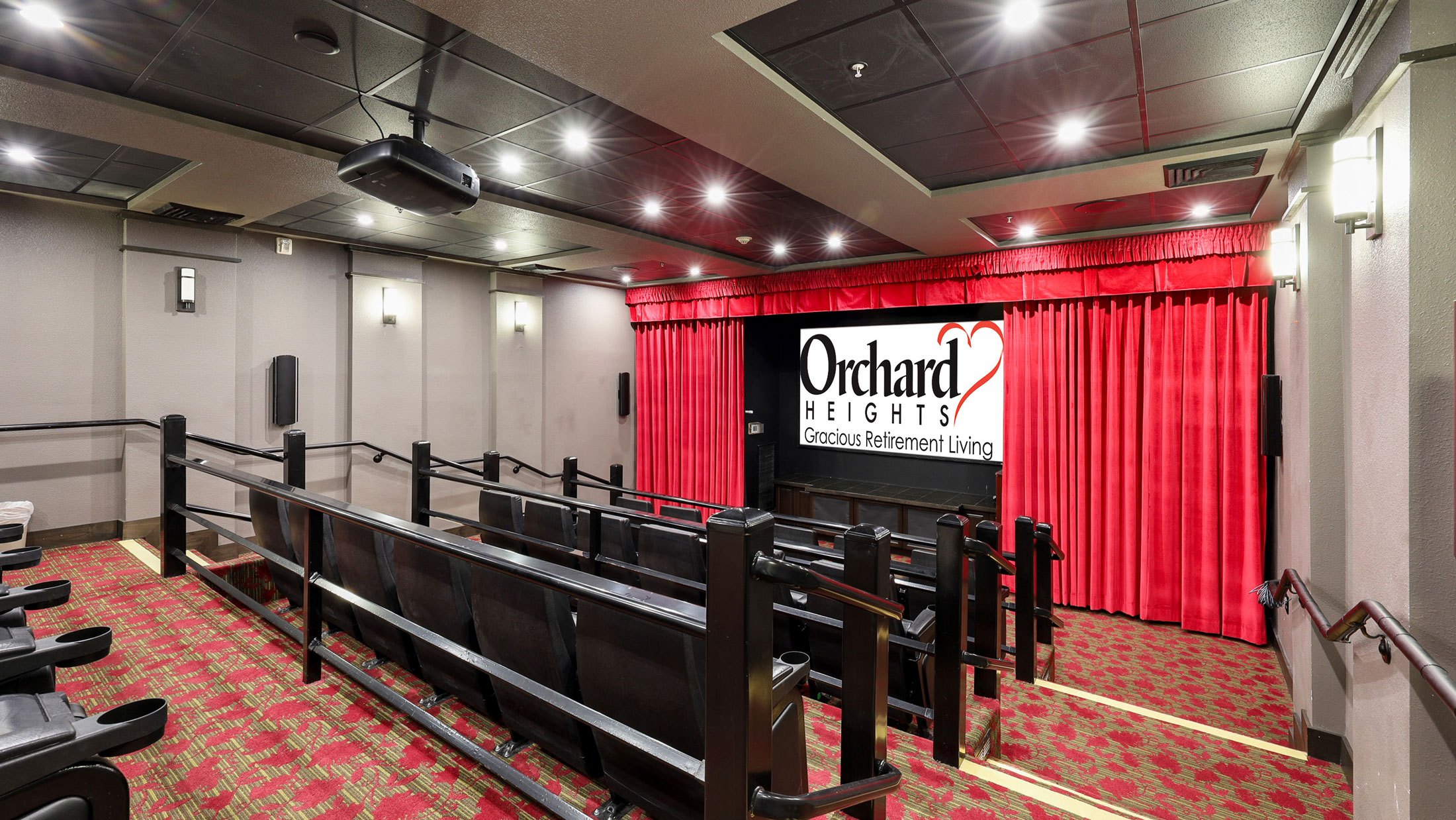 A small movie theater with red carpet, gray walls, and rows of black seats faces a screen displaying the Orchard Heights Gracious Retirement Living logo, with red curtains framing the screen.