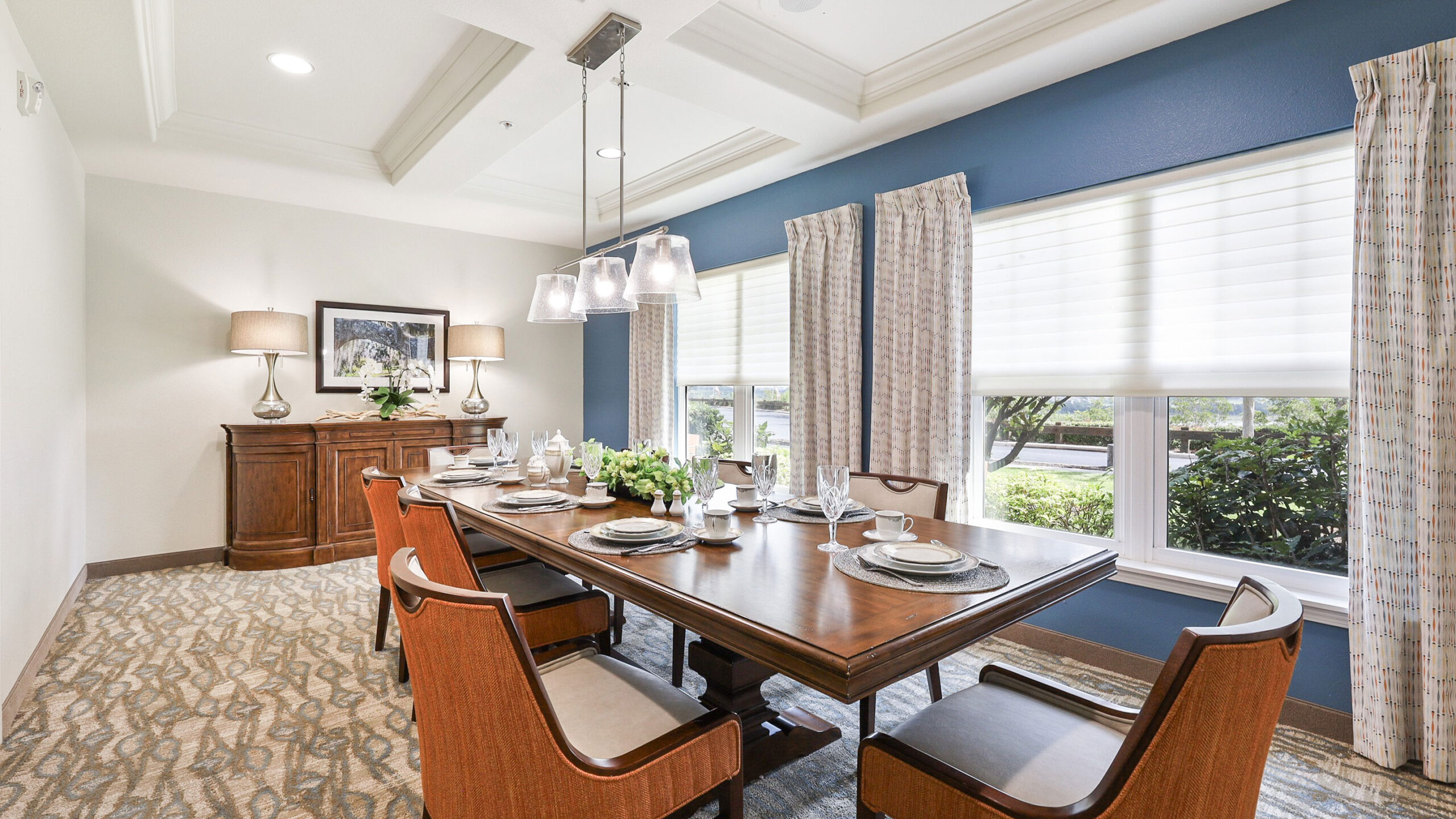 A bright dining room with a wooden table set for six, surrounded by cushioned chairs. Large windows with curtains allow natural light in. A sideboard with lamps and artwork decorates the back wall.