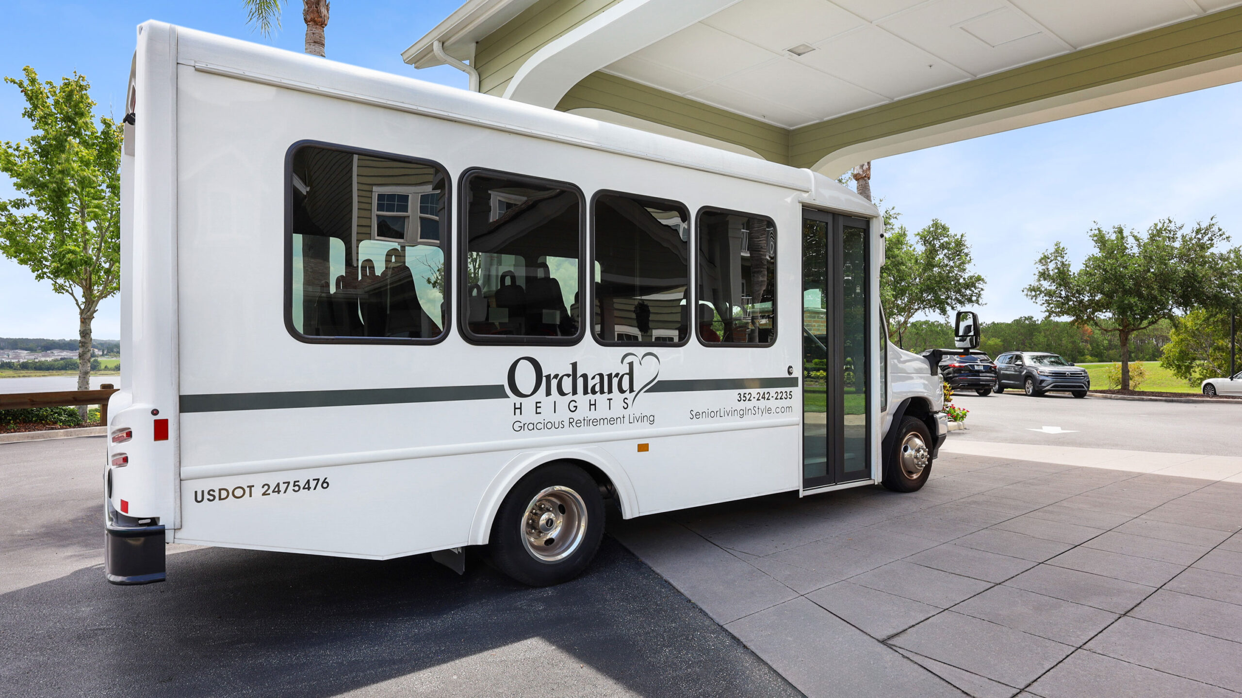 A white shuttle bus with “Orchard Heights Gracious Retirement Living” written on the side is parked outside a building entrance on a sunny day. Trees and a landscaped area are visible in the background.
