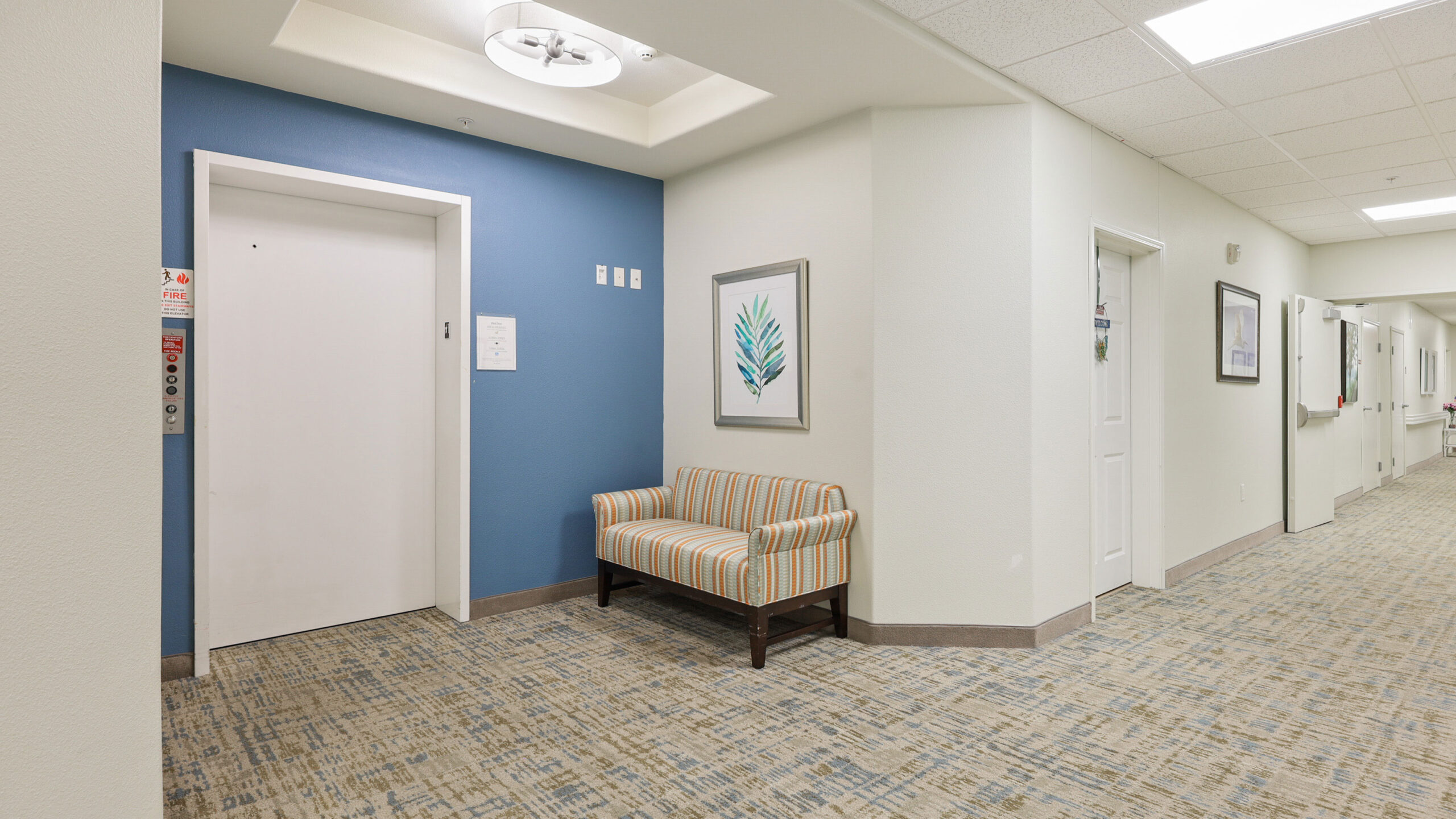 A hallway with patterned carpet, a blue accent wall, an elevator door, a striped cushioned bench, framed artwork, and several white doors leading to rooms. The space is brightly lit with ceiling lights.