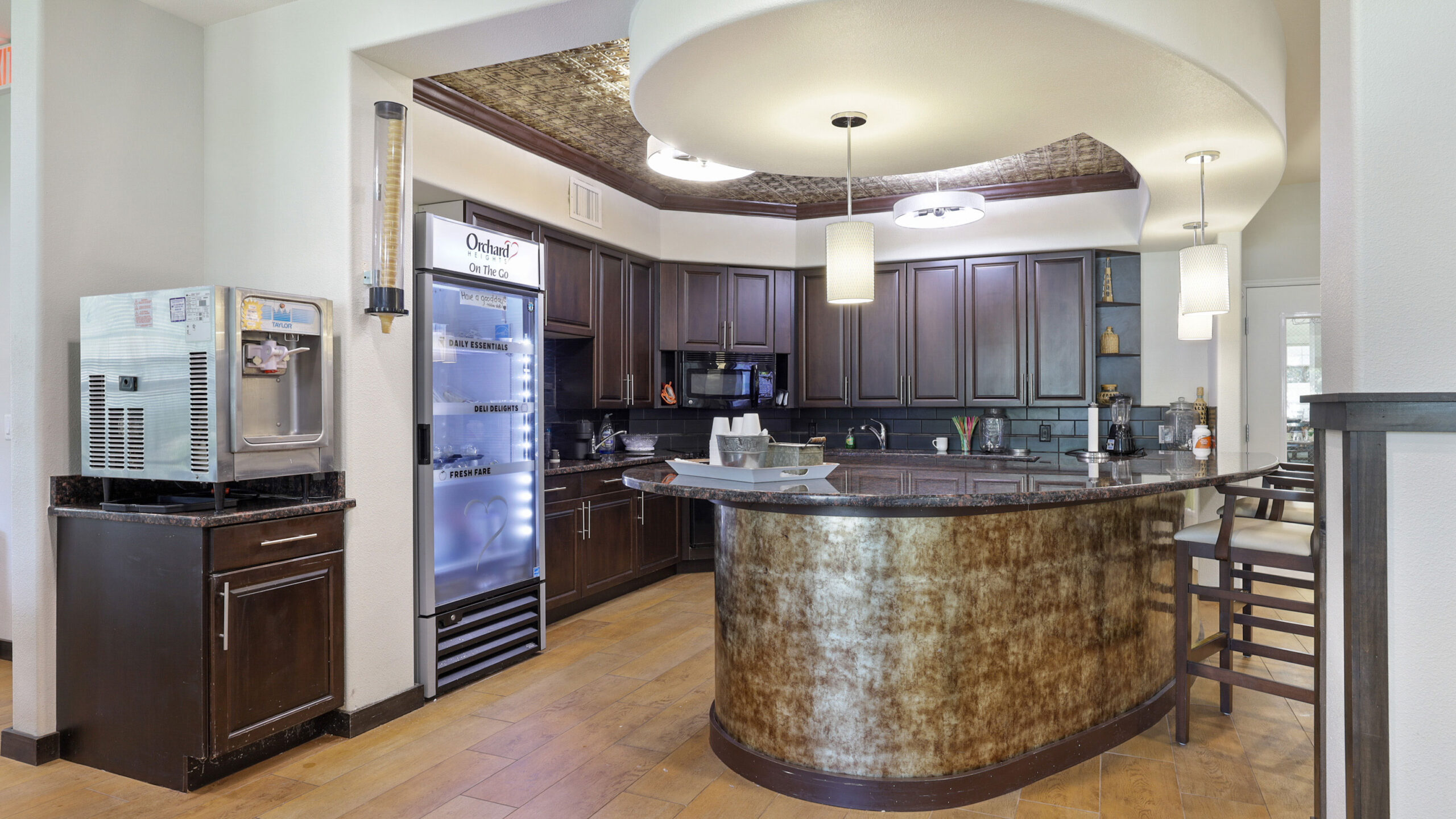 Modern coffee station with dark wood cabinets, a curved granite counter, beverage fridge, ice machine, and pendant lights, set on wood flooring in a brightly-lit area.
