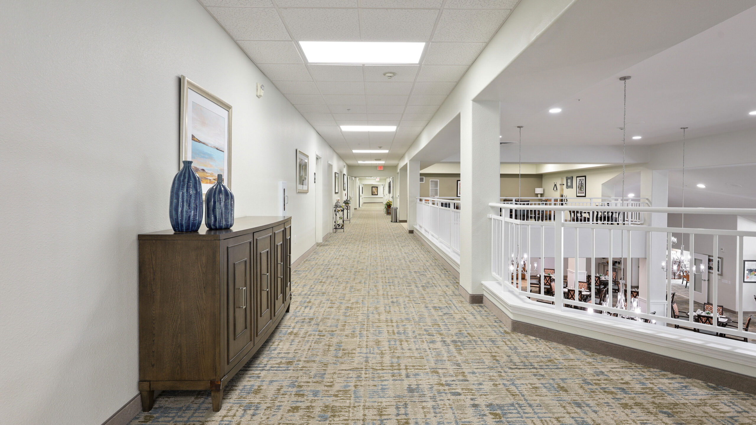 A bright hallway with beige carpet, white walls, a wooden cabinet topped with two blue vases, and framed artwork. The hallway has ceiling lights and overlooks a lower level with white railings.