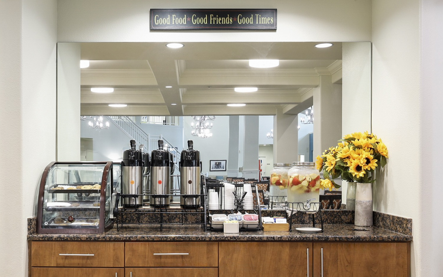 A breakfast buffet counter with coffee dispensers, cups, pastries in a display case, jars of infused water, condiments, and a vase of sunflowers, set in front of a large mirror reflecting a dining area.