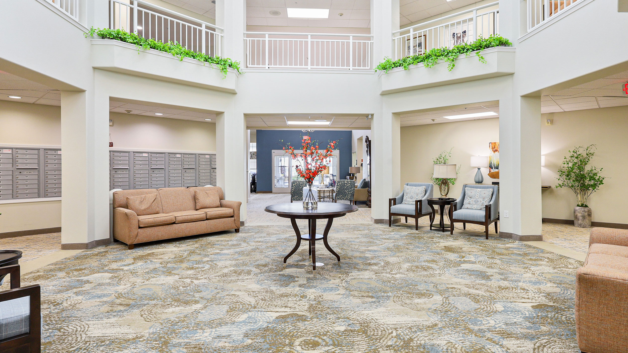 A spacious, well-lit lobby with neutral-toned sofas and chairs, a round table with a flower arrangement in the center, mailboxes on the left wall, and decorative plants on the upper balcony railings.