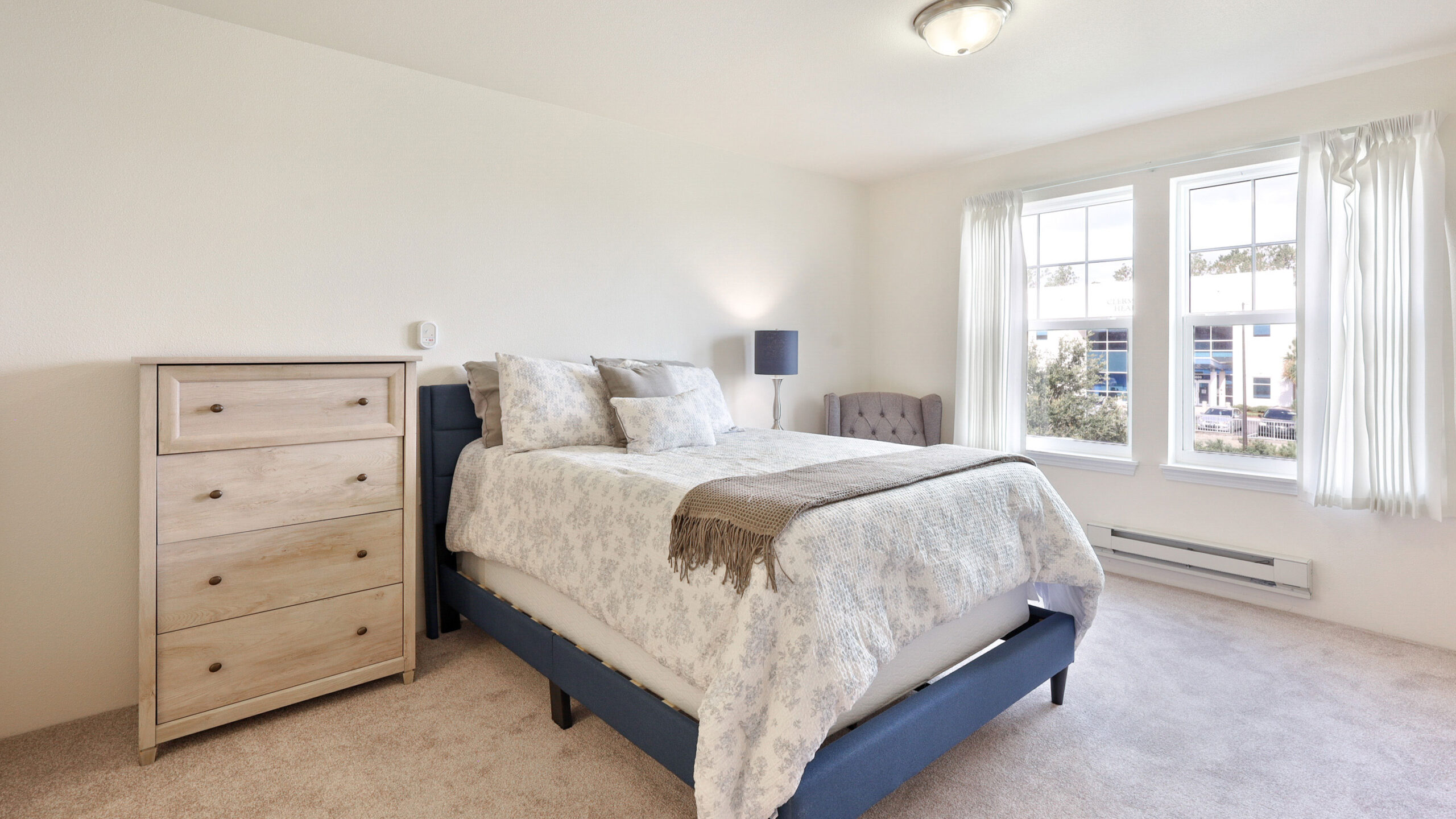 A bright, minimalist bedroom with a bed dressed in light patterned bedding, a beige dresser, a gray armchair, and large windows with white curtains letting in natural light.