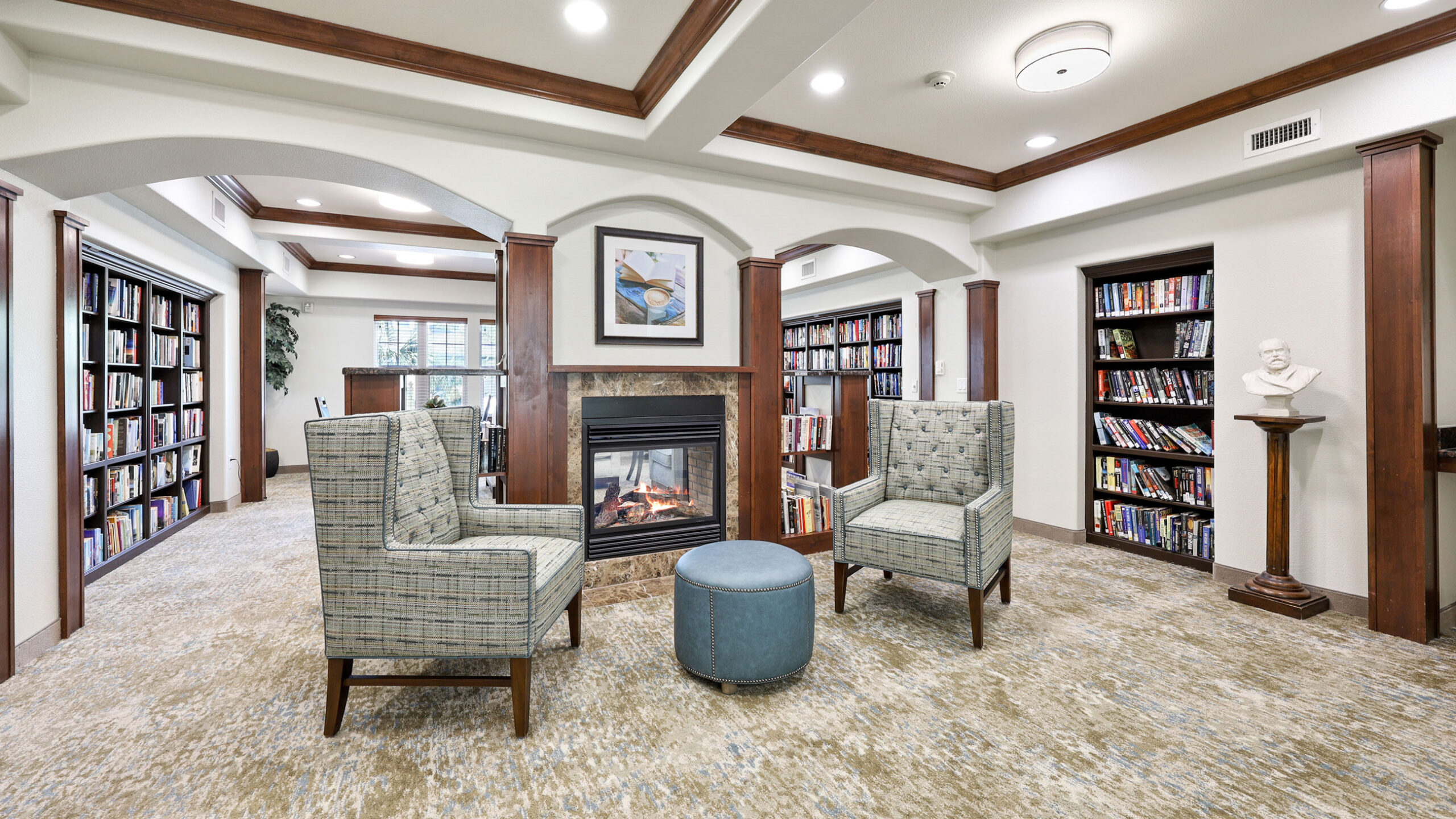 A cozy library with two upholstered chairs and a round ottoman facing a fireplace. Bookshelves line the walls, and a white bust sculpture is displayed on a pedestal near the shelves.