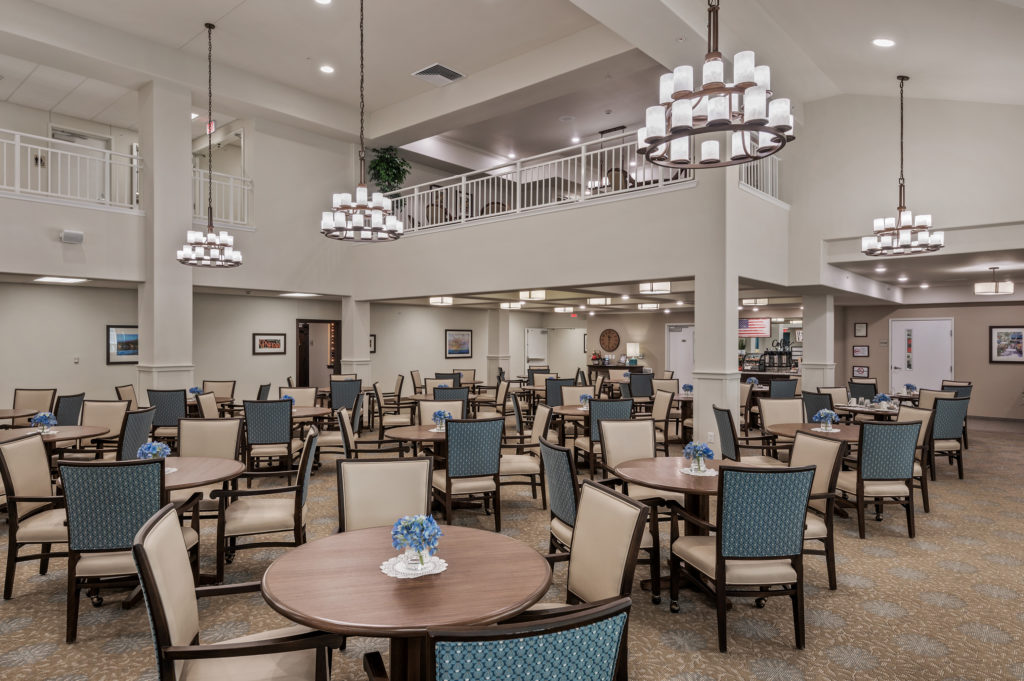 A spacious dining area with high ceilings in a modern facility, featuring round wooden tables with blue and beige chairs. Each table is decorated with a small bouquet of blue flowers. The room is well-lit with large, elegant chandeliers and has a mezzanine level above.