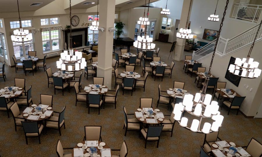 A spacious dining area with multiple round tables covered with white tablecloths and set for meals. Chairs surround the tables, and large chandeliers hang from the ceiling. Windows and a staircase are visible, adding to the elegant ambiance.