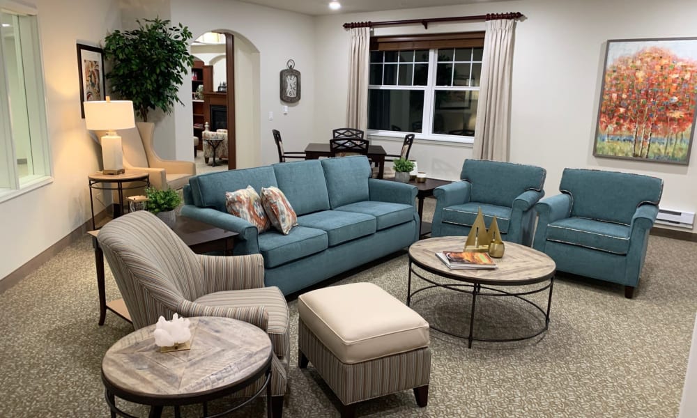 A cozy living room featuring a teal sofa with matching armchairs, a striped armchair with an ottoman, and round wooden coffee tables. The space is decorated with a large tree plant, table lamps, and a colorful painting of autumn trees on the wall.