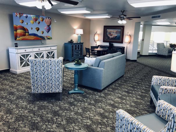 A spacious living room with patterned armchairs, a blue couch, and a round side table. The room features a TV mounted on the wall showing colorful hot air balloons, along with a prominent painting. The space is well-lit with lamps and ceiling lights.