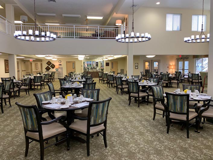 A spacious dining area with multiple round tables covered in white tablecloths and set for meals, featuring cushioned chairs. The room is well-lit with large chandeliers, and a balcony overlooks the dining space. Windows and light beige walls add to the airy ambiance.