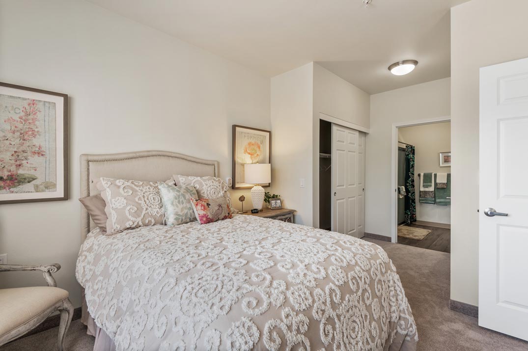 A cozy bedroom with a queen-sized bed featuring a light-colored, intricately patterned comforter and multiple decorative pillows. The room has light walls, framed artwork, a nightstand with a lamp and books, and an open door leading to a bathroom.