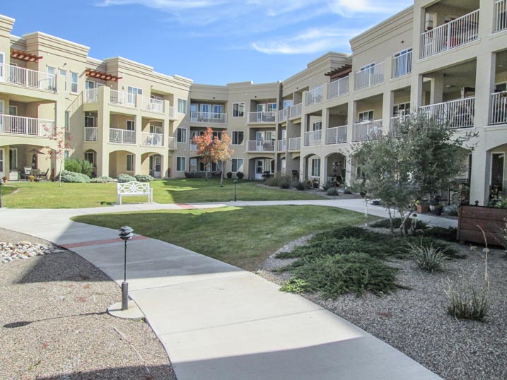 A modern, beige-colored three-story apartment complex with multiple balconies surrounding an inner courtyard. The courtyard features green lawns, small shrubs, pathways, and a few benches. The sky is clear with scattered clouds.